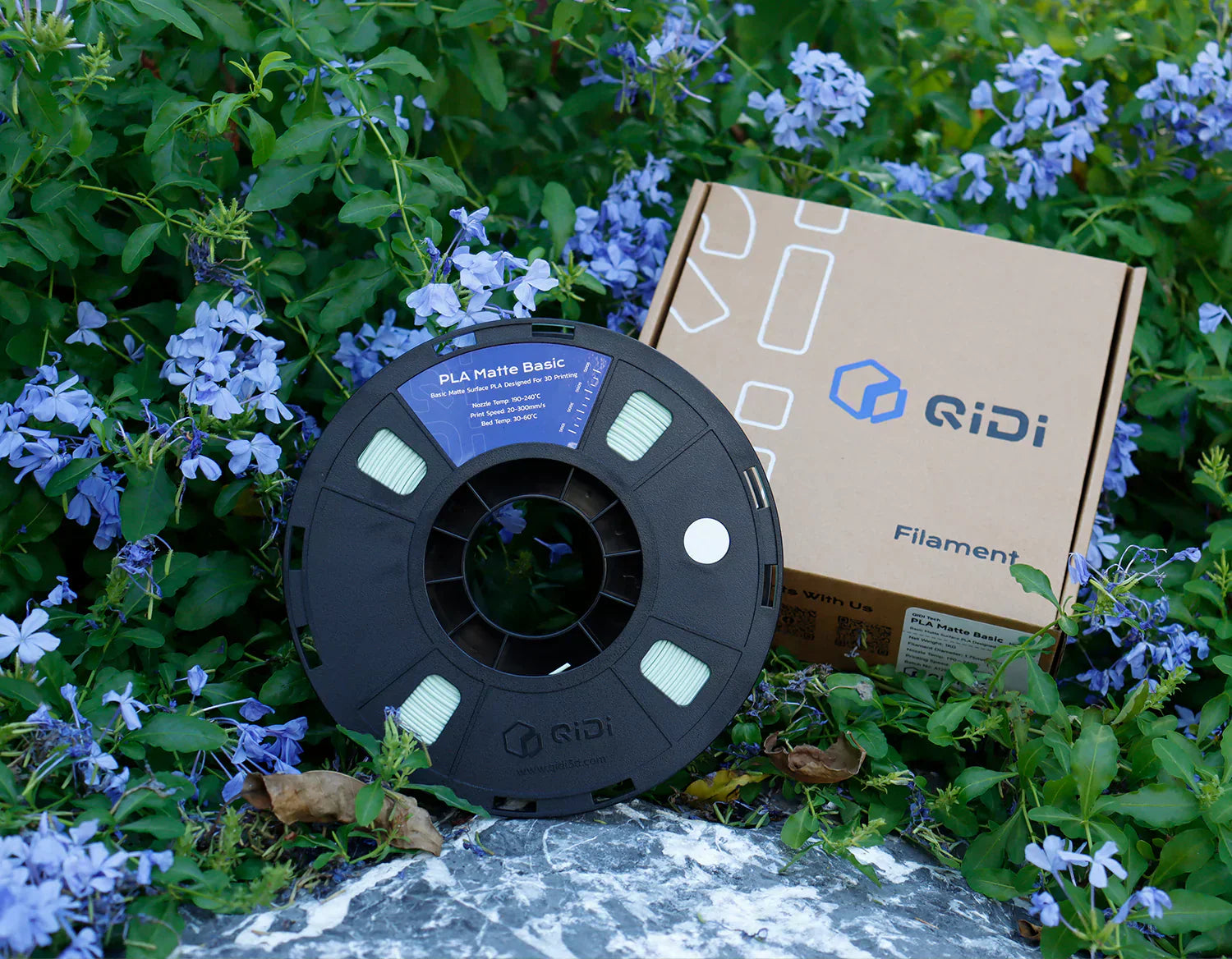 QIDI PLA 3D printer filament spool with branded box, displayed outdoors among purple flowers.